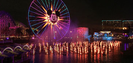 Colorful Ferris wheel with Mickey Mouse face and water fountain show at Disneyland at night.